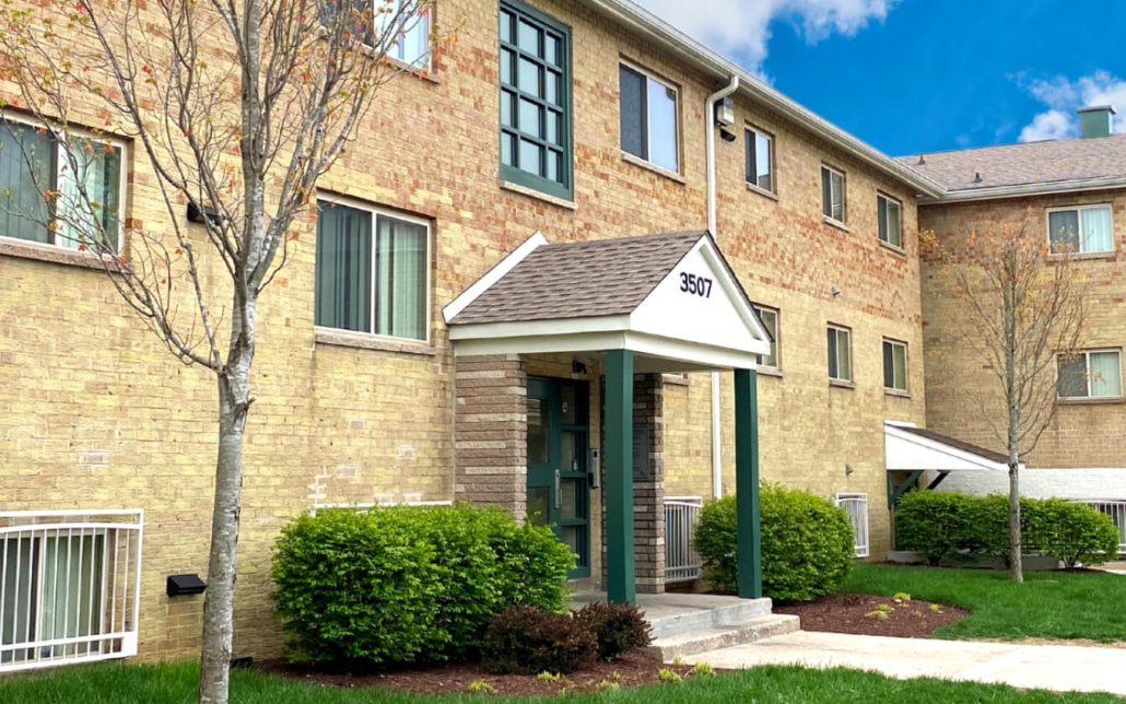 Two-story brick apartment building with a white-trimmed entrance labeled 3507, green doors and windows, small shrubs, and neatly landscaped grass under a partly cloudy sky.