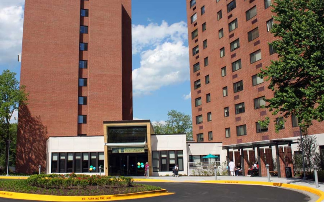 Two tall red-brick apartment buildings are connected by a white entrance with glass doors. People are seen near the entrance, and trees are visible around the buildings under a partly cloudy sky.