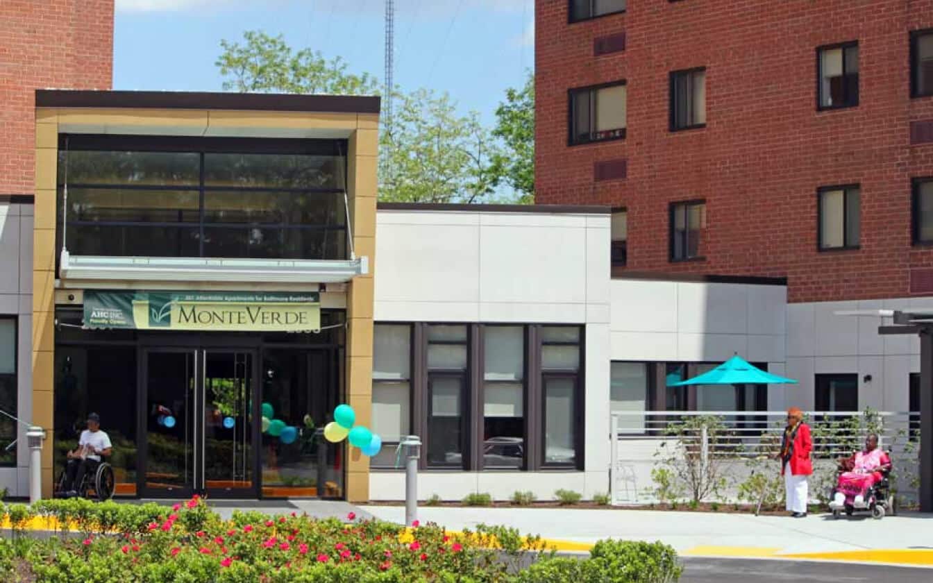 The entrance to MonteVerde, a residential building with large windows. Two people, one in a wheelchair and one standing, are near the door. Another person pushes a wheelchair by a patio with a blue umbrella. Flowers are in the foreground.