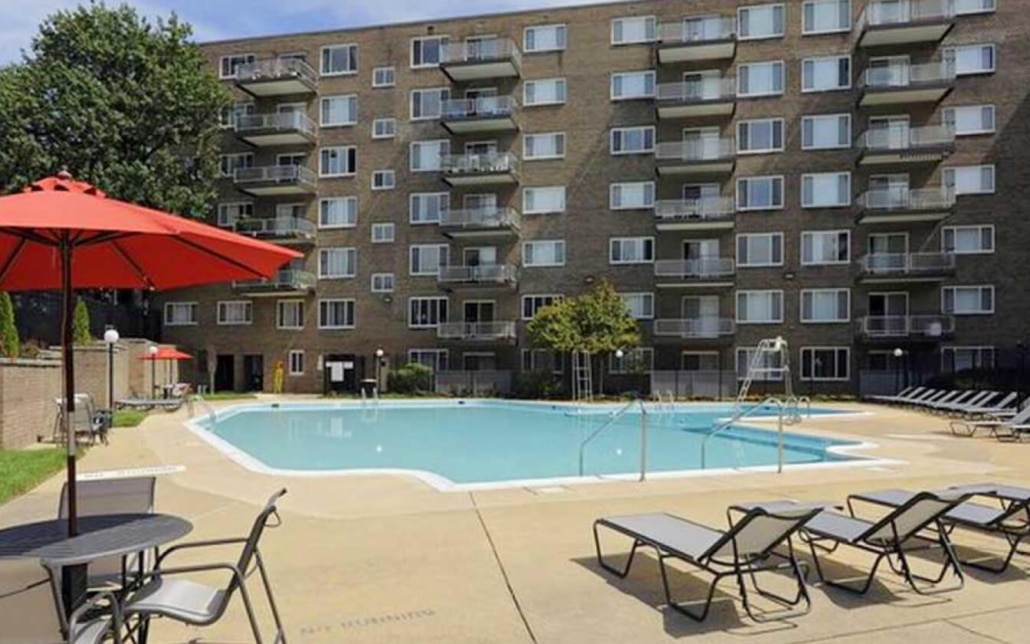 Apartment building with balconies overlooks an outdoor swimming pool surrounded by lounge chairs, tables, and red umbrellas on a sunny day. Trees and fencing border the pool area.