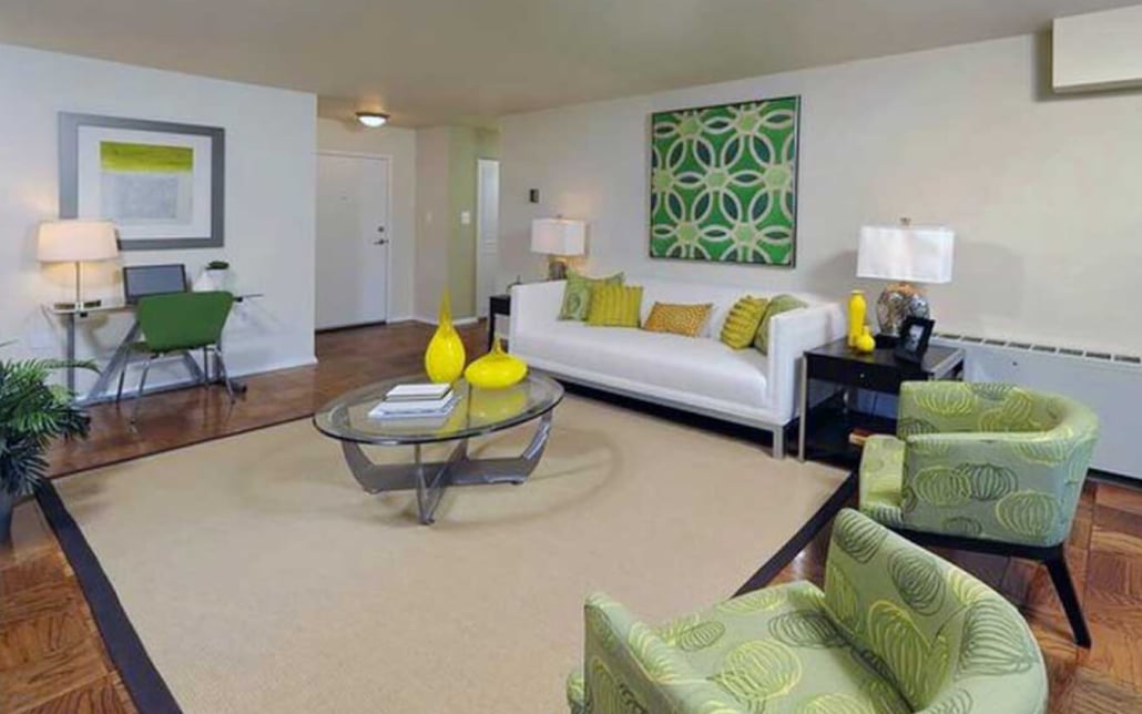 Modern living room with green and yellow accents, featuring a white sofa, two patterned green chairs, a glass coffee table with yellow vases, desk area, and abstract wall art on cream-colored walls and wood flooring.