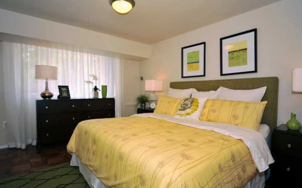 A cozy bedroom with a large bed covered in yellow bedding, two framed pictures above the headboard, dark wood dresser, two bedside tables with lamps, and a window with sheer white curtains letting in natural light.