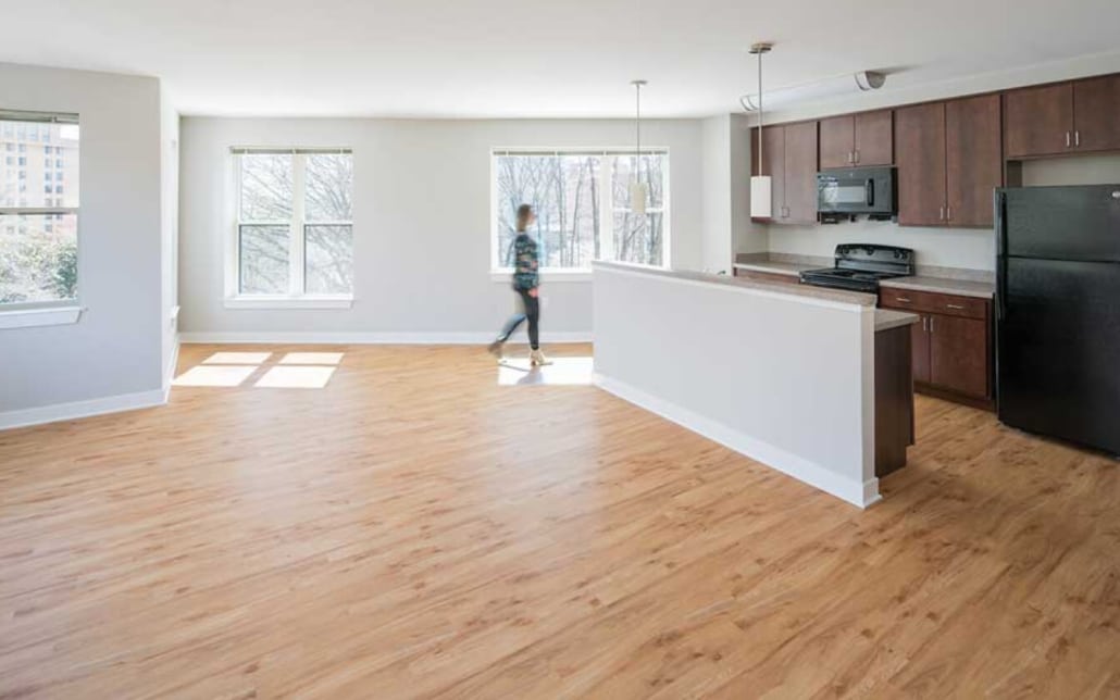 A person walks through a bright, empty apartment with large windows, wood flooring, and an open kitchen with dark cabinets and black appliances.