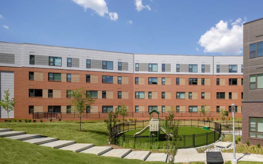 A modern four-story apartment building with red brick and light gray siding surrounds a small fenced playground with a slide and green lawn under a blue sky with a few clouds.