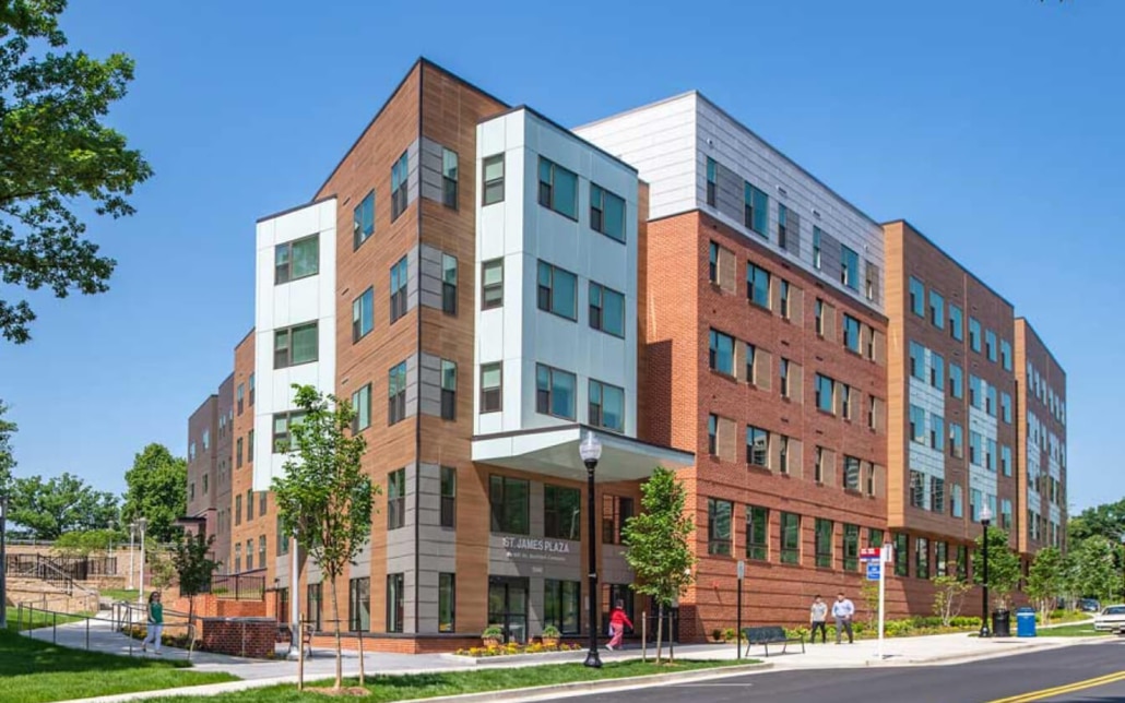 A modern, multi-story apartment building with a mix of brick, wood, and white panel exteriors stands on a sunny day. Trees, a sidewalk, and people walking are visible in front.