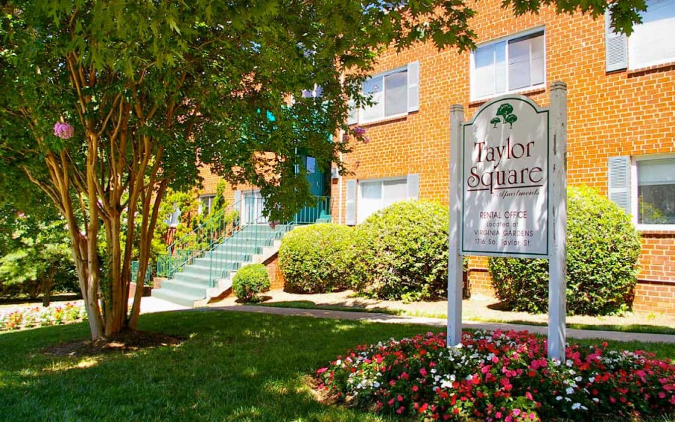 A brick apartment building with green stair railings, surrounded by shrubs and a large tree. In the foreground, a white sign reads “Taylor Square Apartments” amid flowers and manicured grass.