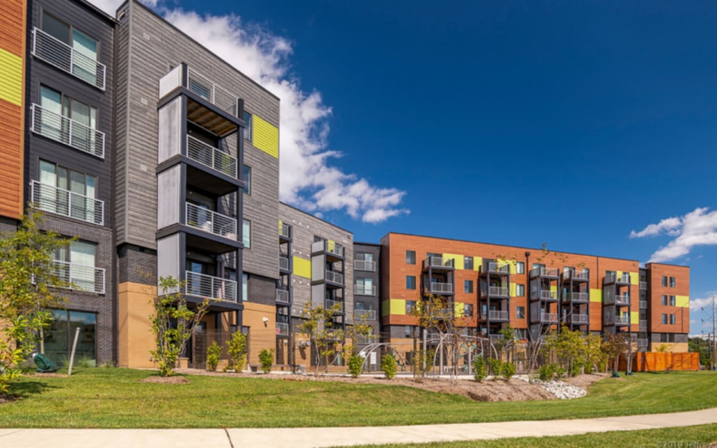 Modern apartment complex with four-story buildings featuring balconies, colorful panels, and large windows, surrounded by green lawns, young trees, and a clear blue sky.