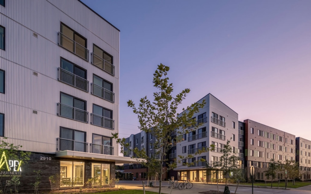 Modern apartment buildings at dusk with lit windows, balconies, and young trees lining the sidewalk. A sign reads APEX Apartments and a bicycle is parked near the entrance. The sky is clear with a soft purple hue.