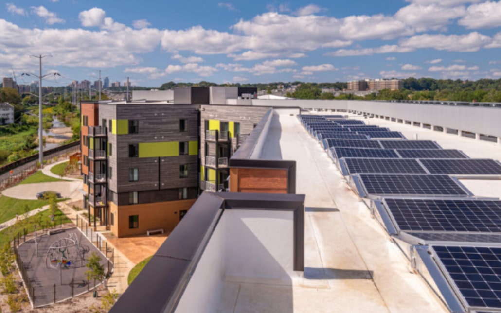 A rooftop covered with solar panels overlooks a modern apartment building, playground, and green landscaped area under a blue sky with scattered clouds.