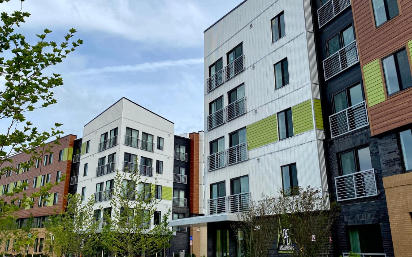 Modern apartment buildings with varied facades in white, green, brown, and black tones, featuring large windows and balconies. Young trees line the foreground under a partly cloudy sky.