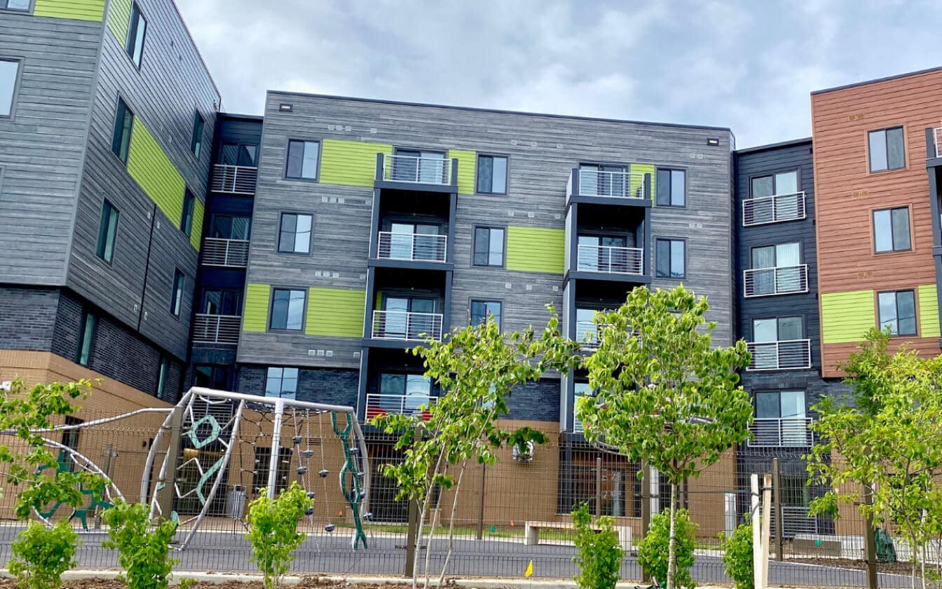 Modern apartment building with green and brown panels, multiple balconies, and large windows. In front, there is a small playground with climbing equipment and young trees growing along a fence. The sky is cloudy.