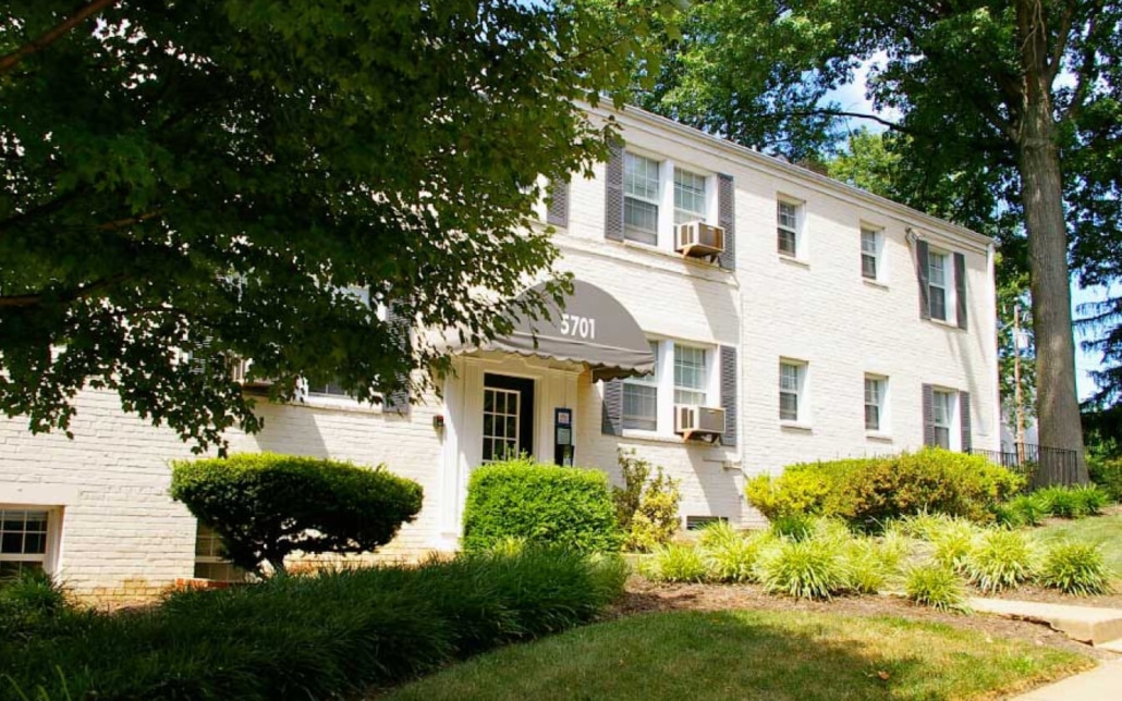 A two-story white brick apartment building with gray shutters and window air conditioners, surrounded by green bushes, grass, and large trees under a clear sky. The address 5701 is on a black awning above the entrance.