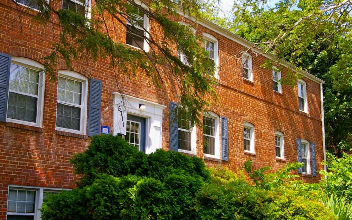 A two-story brick apartment building with white-trimmed windows, blue shutters, and a white entrance surrounded by lush green bushes and trees on a sunny day.