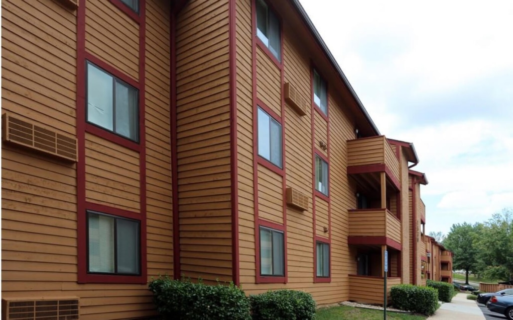 A three-story apartment building with brown wooden siding and red trim, featuring several small balconies, shrubs along the base, and a partly cloudy sky in the background.