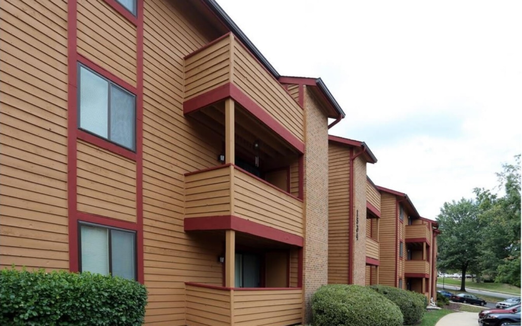 A three-story apartment building with brown wooden siding and red trim, featuring several small balconies. Bushes line the front and cars are parked to the right along the street.