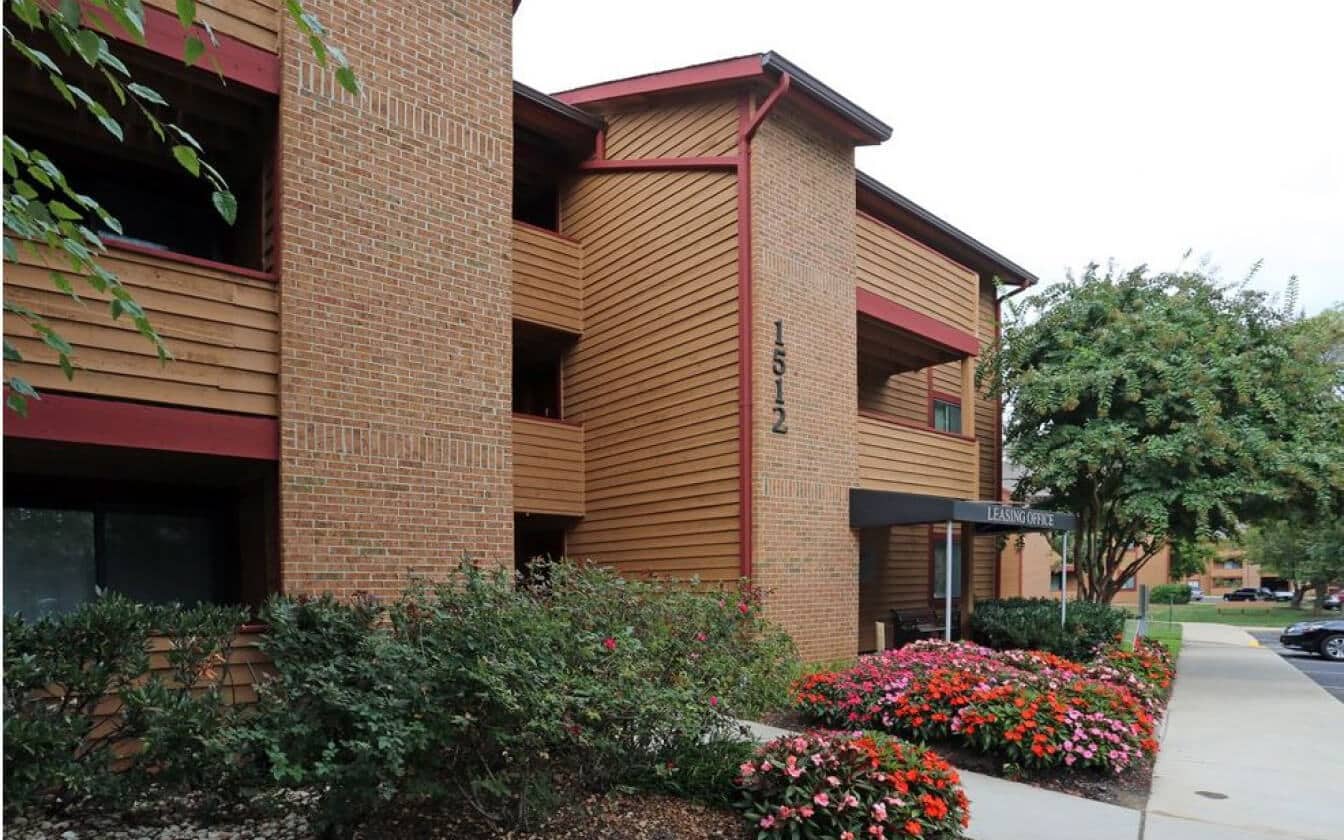 A brick and wood apartment building at 1512, with a covered entrance, colorful flower beds, green shrubs, and a tree-lined sidewalk on a cloudy day.