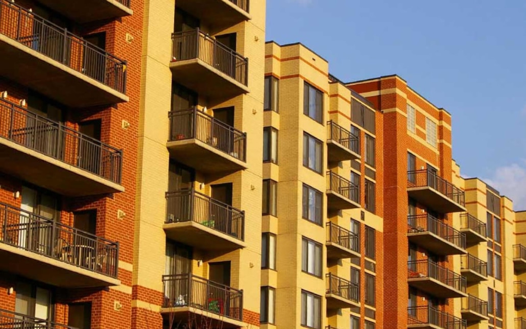 A modern apartment building with multiple floors features balconies on each unit, a mix of tan and red brick exterior, and large windows, all under a clear blue sky.