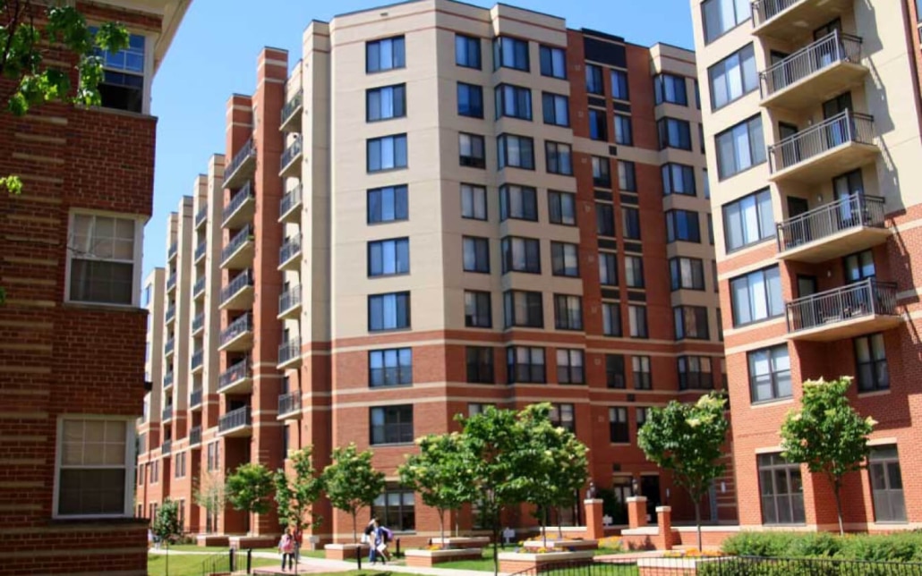 Several mid-rise apartment buildings with brick and beige exteriors surround a courtyard with green trees and walkways, on a sunny day. A few people are visible walking near the entrance.