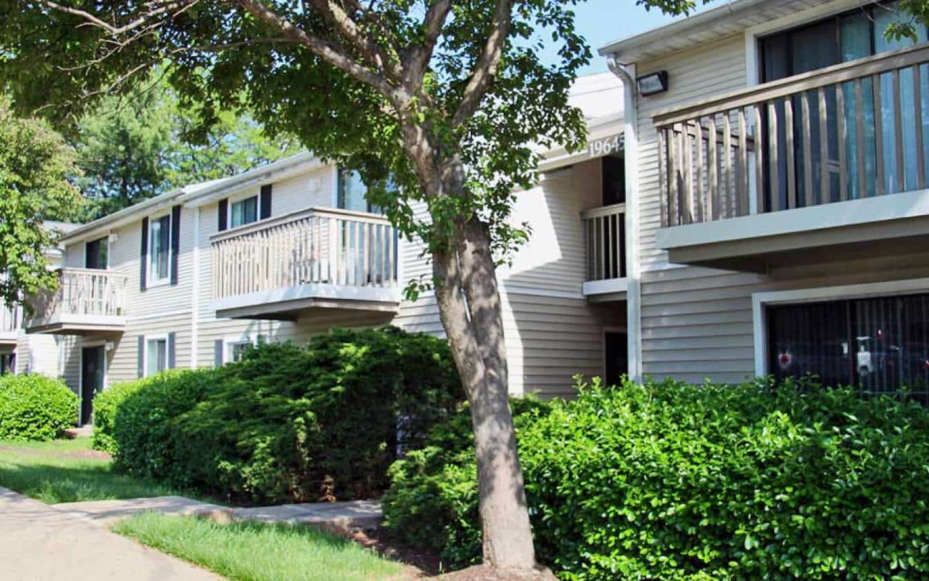 Two-story apartment building with beige siding, several small balconies, and large windows, surrounded by green bushes, trees, and a sidewalk in a landscaped outdoor area on a sunny day.