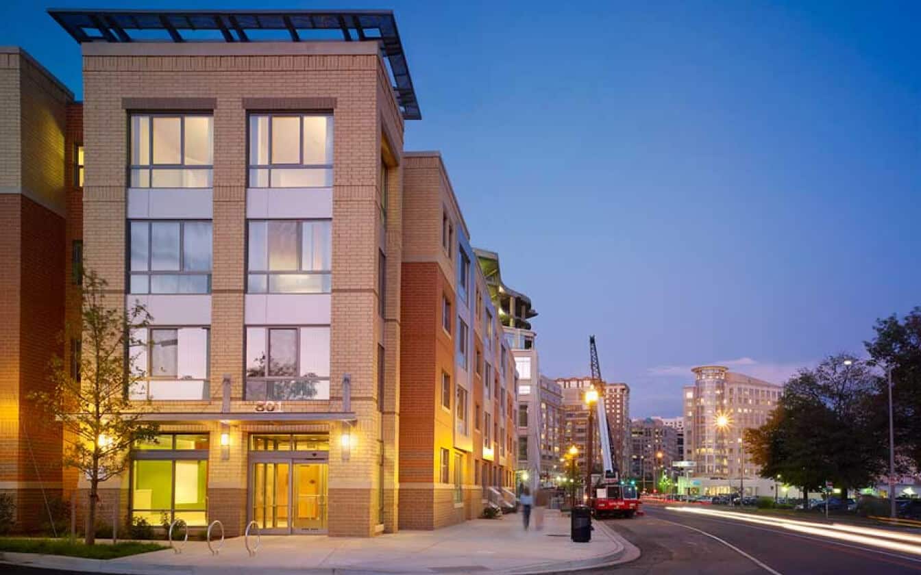 A modern apartment building with large windows and warm lighting stands at a city street corner at dusk. Construction equipment is visible down the street, and other buildings are illuminated in the background.