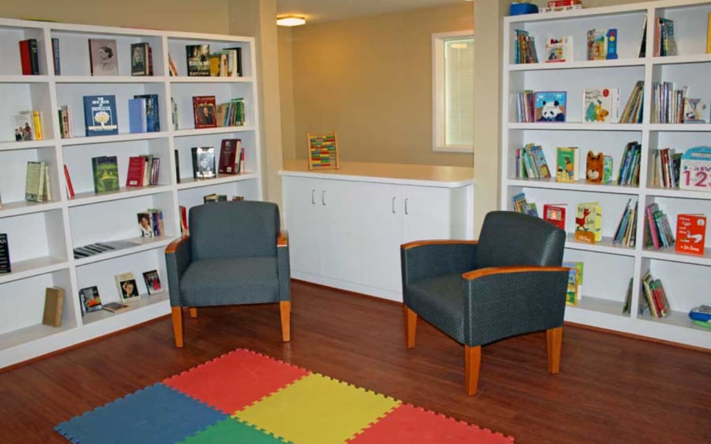 A cozy reading room with two gray armchairs, white bookshelves filled with books, a white cabinet, and a colorful foam mat on a wood floor. A window and a wooden bead maze toy are in the background.