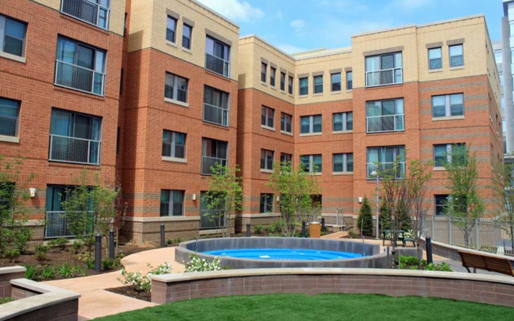 A landscaped courtyard surrounded by a multi-story brick apartment building, featuring a round fountain in the center, garden beds, young trees, benches, and a well-maintained grassy area.