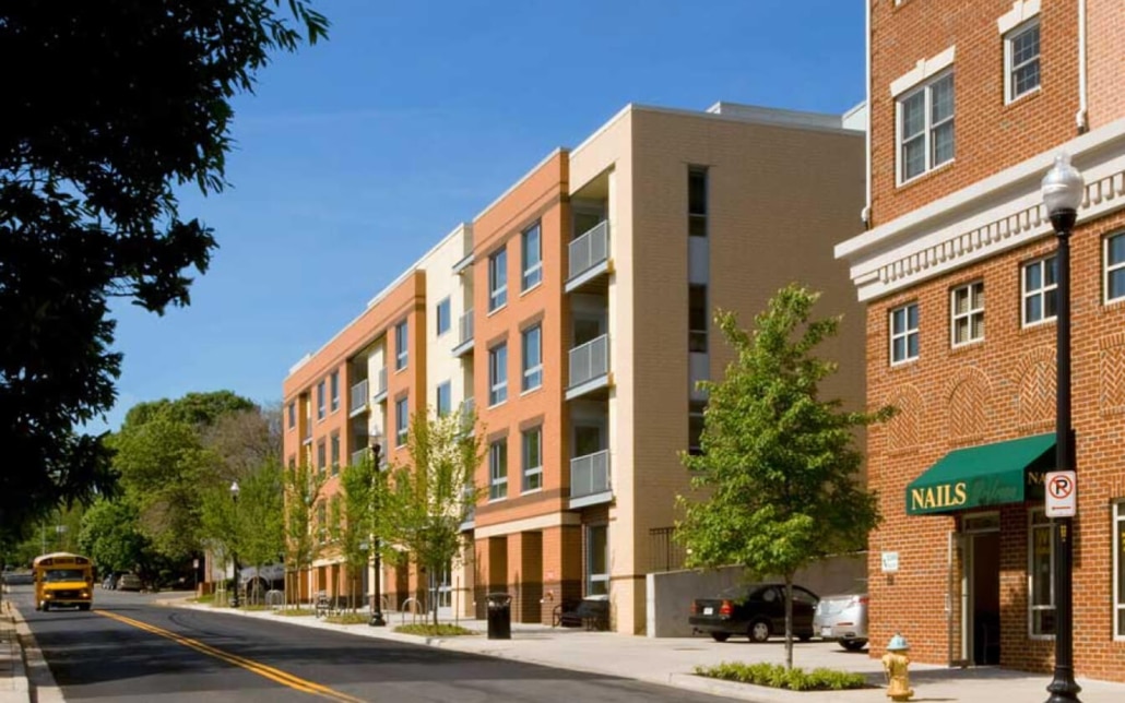 A tree-lined street with modern brick apartment buildings, a sidewalk, and a small business called “Nails.” A yellow school bus is parked in the background under a clear blue sky.