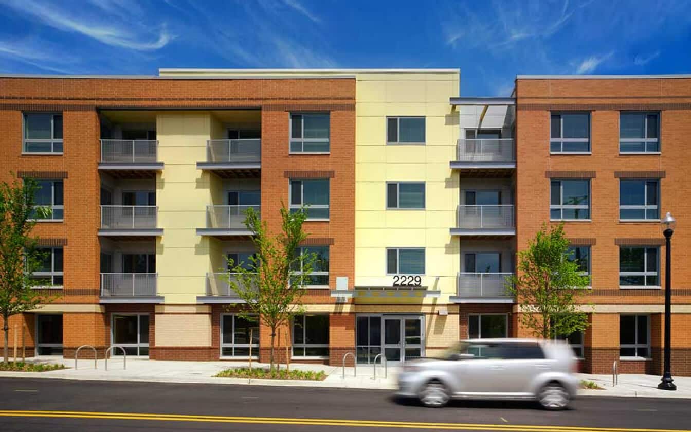 A modern three-story apartment building with yellow and brown brick facade, balconies, and large windows; a white car is driving past on the street in front.