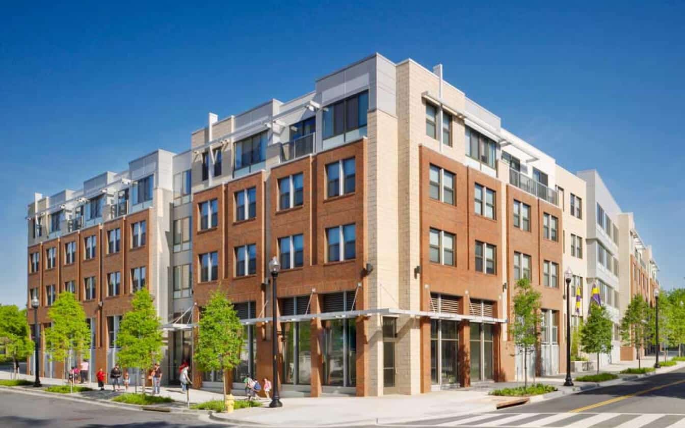 A modern, multi-story apartment building with red brick and light-colored accents stands on a sunny street corner, surrounded by trees and a few pedestrians.