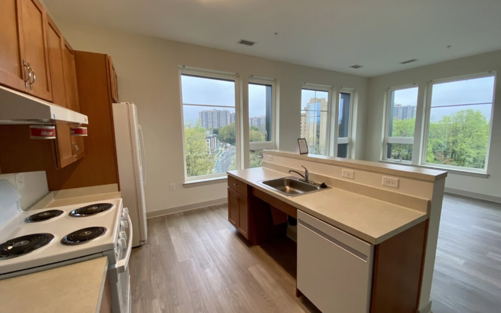 Modern apartment kitchen with light wood floors, white appliances, brown cabinets, and large windows offering a city and greenery view. A sink and countertop island face the open living area.