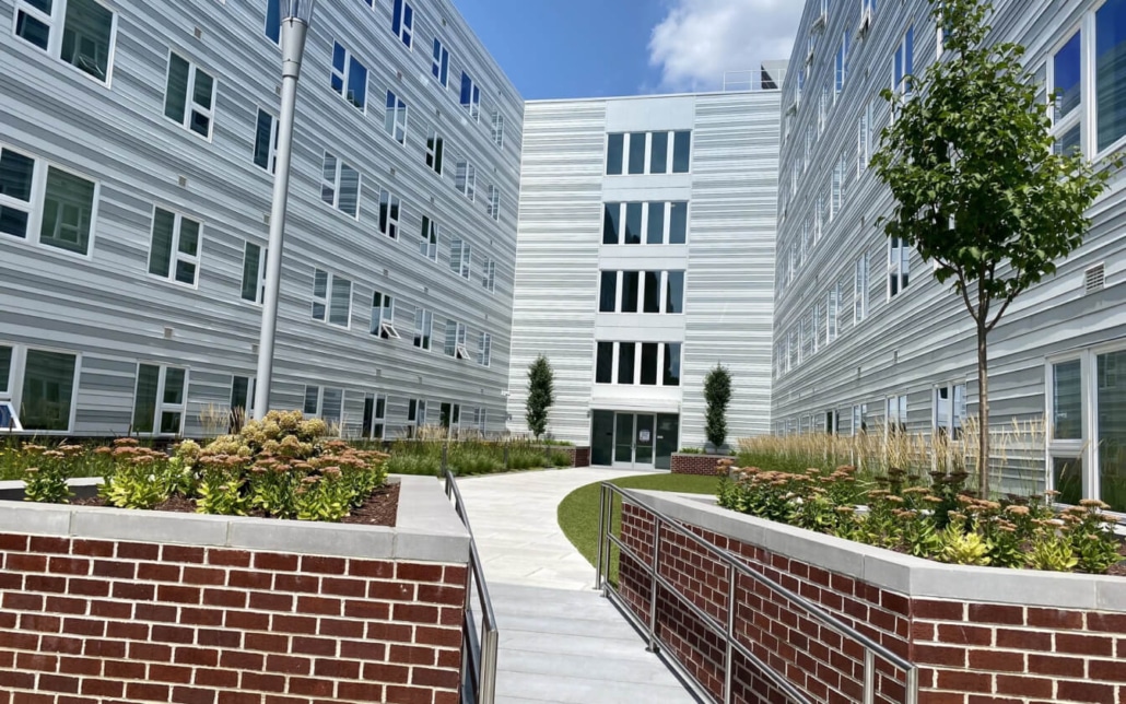 A modern building complex with white and gray exterior walls, large windows, landscaped flower beds, a curved walkway, and a few young trees under a partly cloudy sky.
