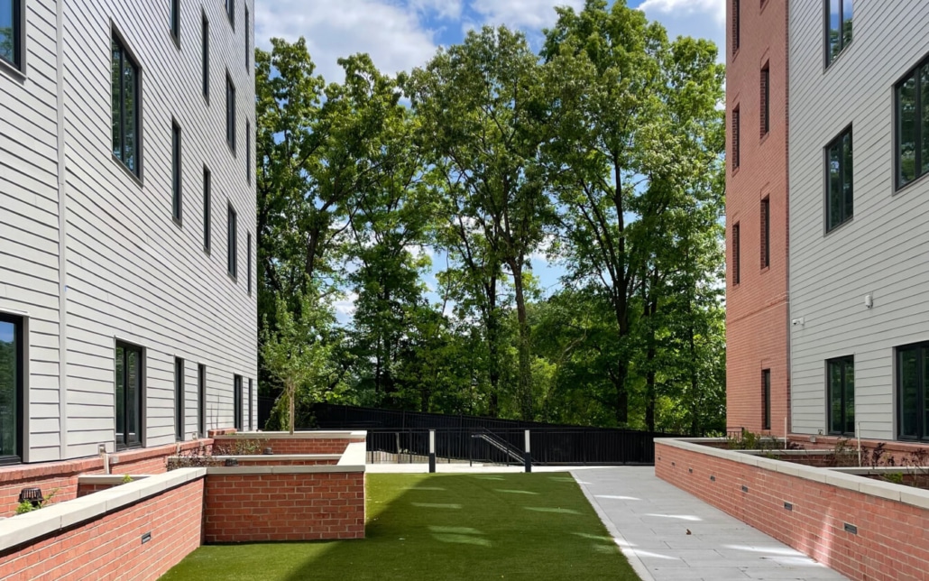 A small courtyard with artificial grass and brick planters is situated between two modern buildings, with trees and greenery visible beyond a black fence under a partly cloudy sky.