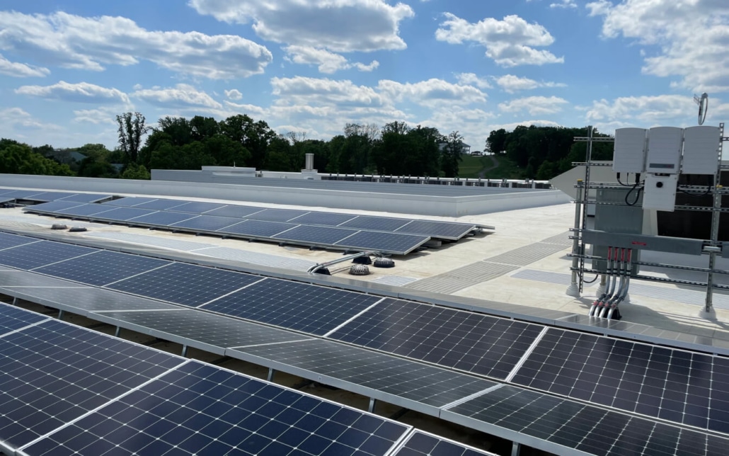 Rows of solar panels are installed on a flat rooftop under a partly cloudy sky, with trees and greenery visible in the background. Electrical equipment and wiring are mounted nearby.