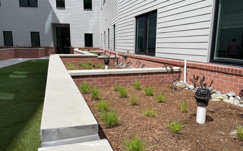 A landscaped area with young green plants and mulch, bordered by brick walls next to a building with gray siding and black windows; pathway and outdoor lights are visible.