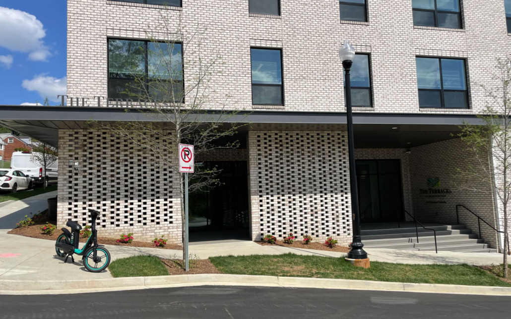A brick building with large windows and a covered entrance is shown. A “No Parking” sign stands near a sidewalk with electric scooters parked beside it. Small plants line the building’s exterior.
