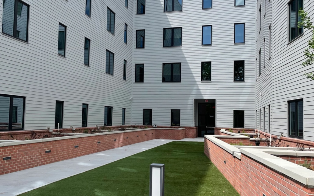 A modern apartment courtyard with artificial grass, brick planters, and a concrete path, surrounded by light gray siding and many windows. There is a door at the far end of the courtyard.