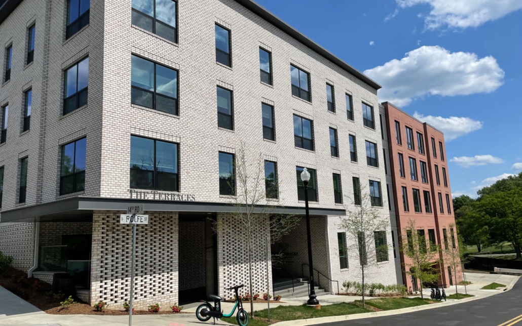 A modern four-story apartment building with light brick and large windows, featuring a shaded entrance, a bike and scooter parked outside, a street sign, and trees and greenery nearby under a blue sky with clouds.