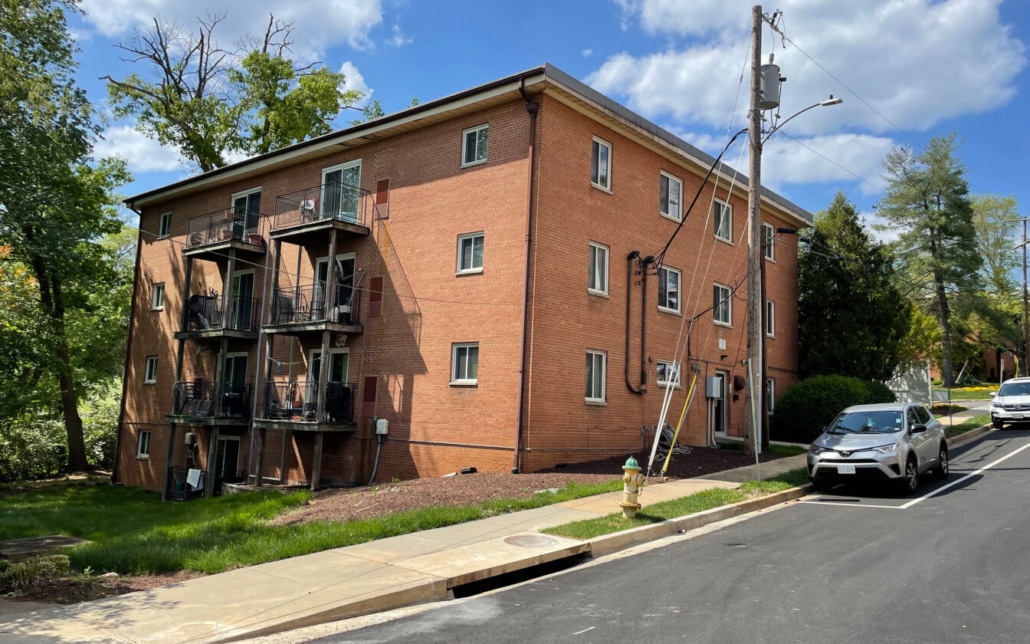 A three-story red brick apartment building with balconies on one side, next to a sidewalk and a sloped street with parked cars, trees, and a fire hydrant in the foreground under a partly cloudy sky.