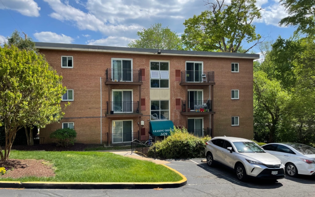 A three-story brick apartment building with balconies, an awning over the entrance, and several parked cars in front, surrounded by green trees and grass under a partly cloudy sky.