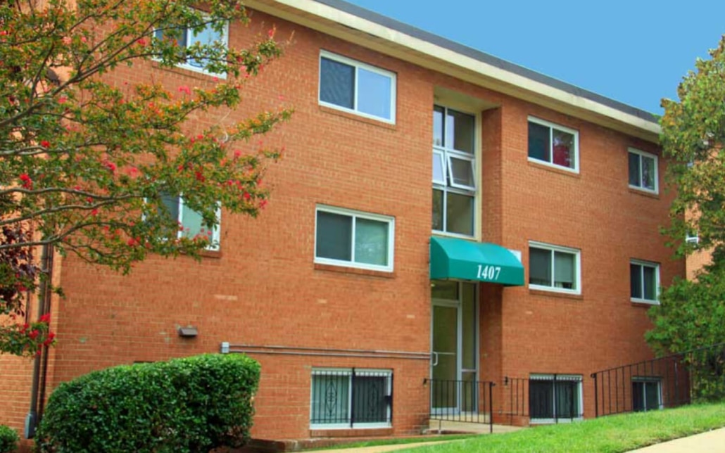 A three-story brick apartment building with white-framed windows and a green awning above the entrance displaying the number 1407. Bushes and trees are in front, and the sky is clear and blue.