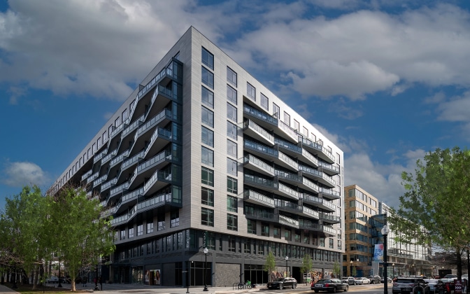 A modern, multi-story apartment building with large balconies and glass windows on a sunny day, surrounded by trees and parked cars along the street.