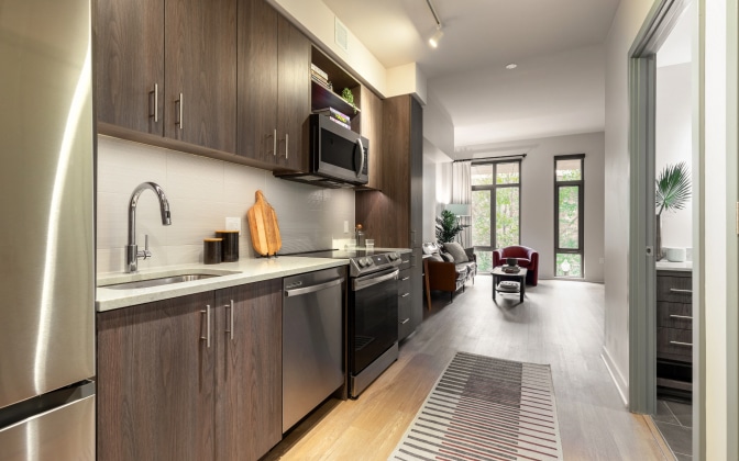 Modern kitchen with dark wood cabinets, stainless steel appliances, and a light countertop. The kitchen opens to a living area with large windows and a sitting area. A patterned rug lines the wooden floor.