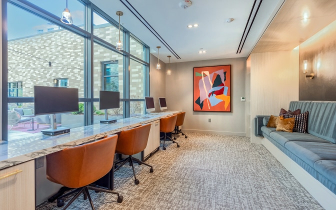 Modern workspace with four computer stations, brown chairs, and a marble countertop. Large windows provide natural light. A colorful abstract painting hangs on the wall above a gray cushioned bench with pillows.