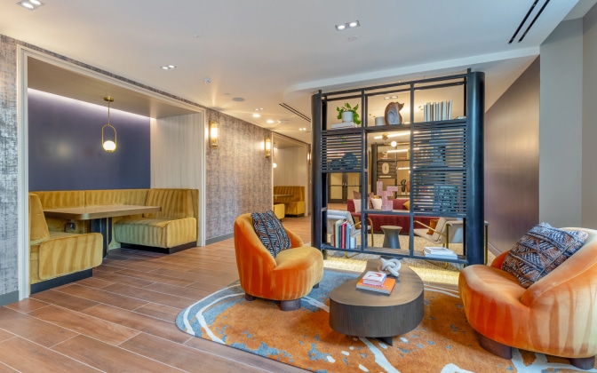 Modern lounge area with two orange velvet chairs, a round wood coffee table on a patterned rug, and two yellow booth seating areas, separated by a decorative black slatted partition. Warm lighting creates a cozy atmosphere.