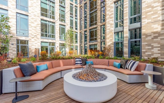 Curved outdoor sofa with cushions and pillows surrounds a modern circular fire pit on a wooden deck, set in a courtyard of a multi-story building with large windows and landscaped plants.