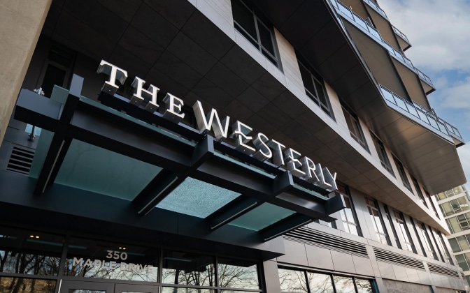 A low-angle view of a modern building entrance with a sign reading THE WESTERLY and the address 350 Maple Drive visible on the glass door beneath a metal awning.