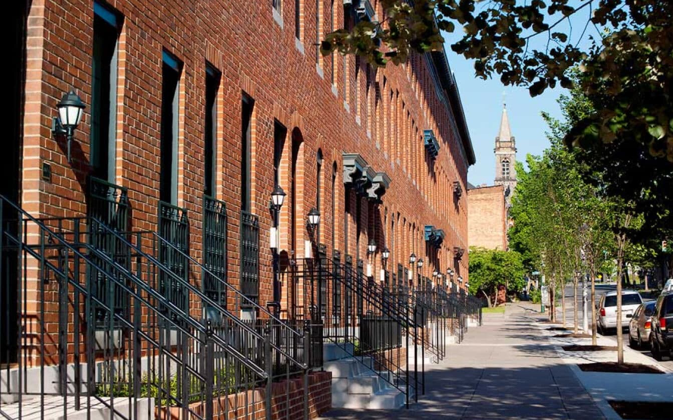 A row of brick townhouses with black railings and steps lines a tree-shaded city sidewalk. Parked cars and a church steeple are visible in the background on a sunny day.