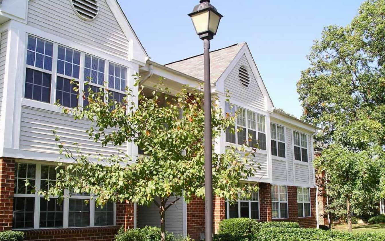 Two-story apartment building with white siding, large windows, and red brick base. A tree and a black lamp post are in front, surrounded by green bushes and grass, under a clear blue sky.