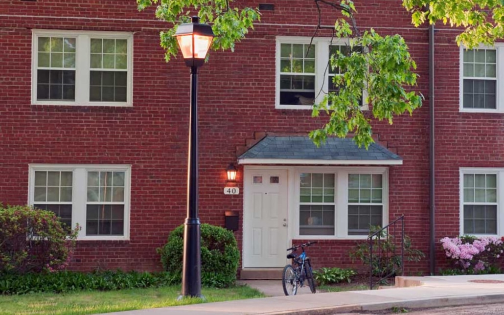 A brick apartment building with white-trimmed windows, a white front door, a black metal lamp post, and a bicycle parked by the entrance. Green trees and bushes line the sidewalk.