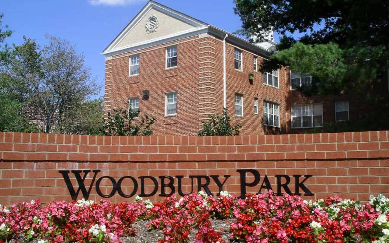 A red brick sign with black letters reading Woodbury Park in front of a flower bed, with a three-story red brick apartment building and trees in the background.